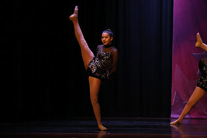 A girl onstage at a dance competition doing a high kick - a grand battement in proprer dance terminology. This is from a jazz dance.