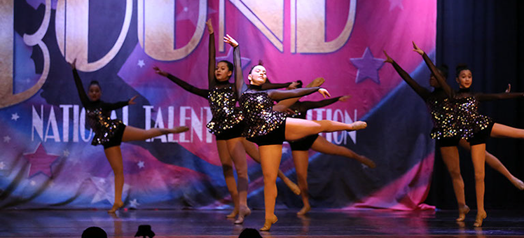 A group of girls onstage at Starbound doing arabesques. They are wearing dark glittery costumes. This picture is from a jazz dance.