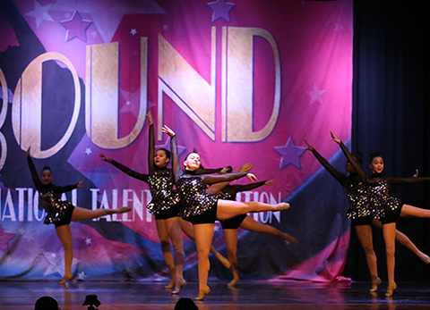 A group of girls onstage at Starbound doing arabesques. They are wearing dark glittery costumes. This picture is from a jazz dance.
