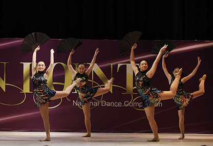 Four girls on stage at OnPoint Dance Competition in "oilslick" costumes and holding fans, doing an attitude. This picture is from a jazz dance.