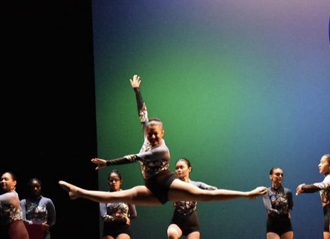 Various girls on stage at Kean University in purple and black costume. One girl is midair doing a perfect leap.